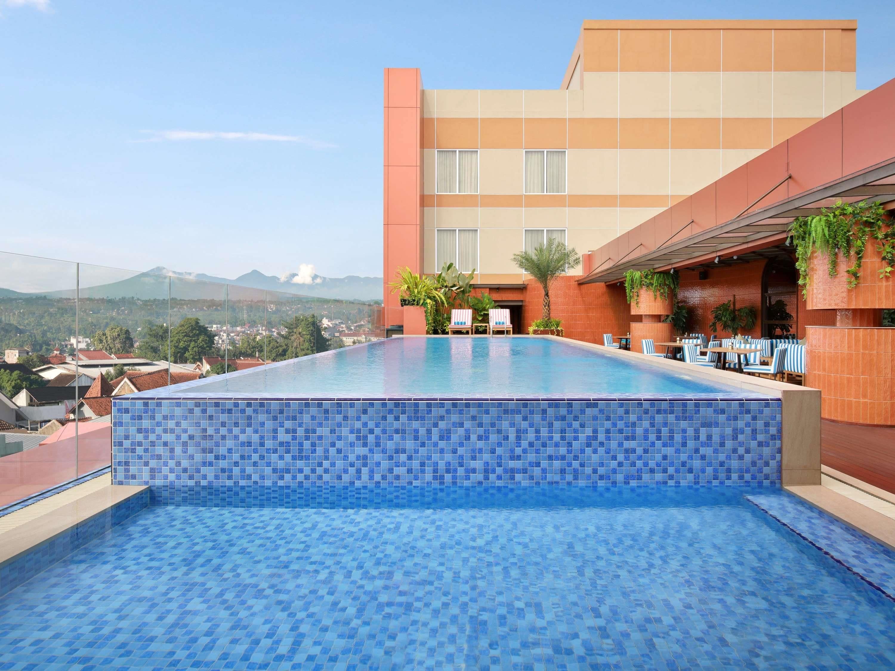 A rooftop infinity pool with a view of distant mountains, a terrace with seating, and a building with a tiled facade.