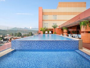 Ein Dachpool mit unendlicher Optik und Blick auf ferne Berge, eine Terrasse mit Sitzgelegenheiten und ein Gebäude mit Kachelfassade.