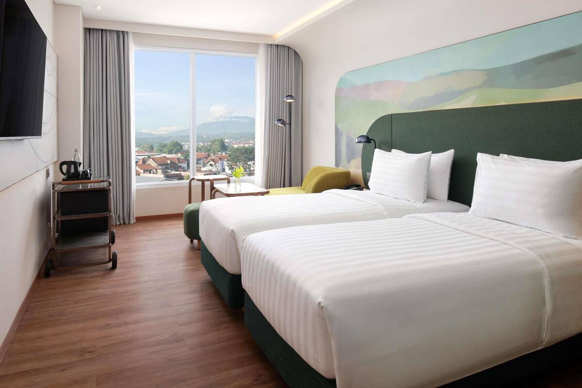 A hotel room with two beds, a desk, a chair, a TV, and a view of the city.