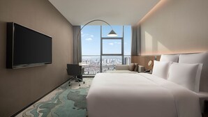 A hotel room with a large bed, a desk, a chair, a flat-screen TV, and a city view through a large window. - Novotel Lianyungang Haizhou (Lianyungang)