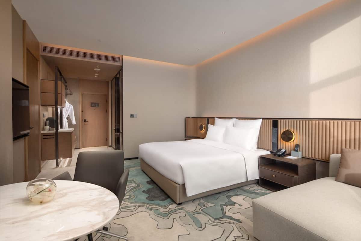Ein modernes Hotelzimmer mit Bett, Nachttisch, Sessel, Schreibtisch und einem kleinen Tisch mit einer Vase.