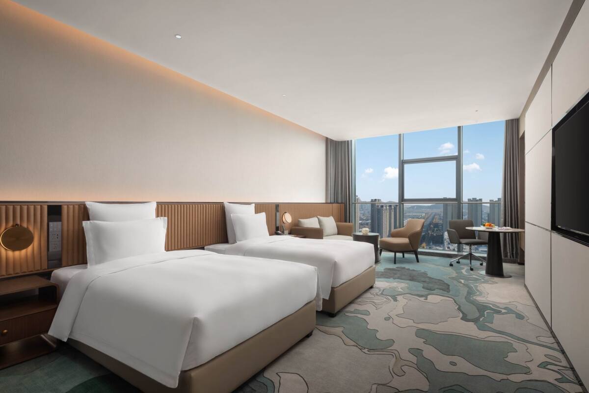 Ein Hotelzimmer mit zwei Betten, einem Schreibtisch, einem Stuhl und einem großen Fenster mit Blick auf die Stadt.