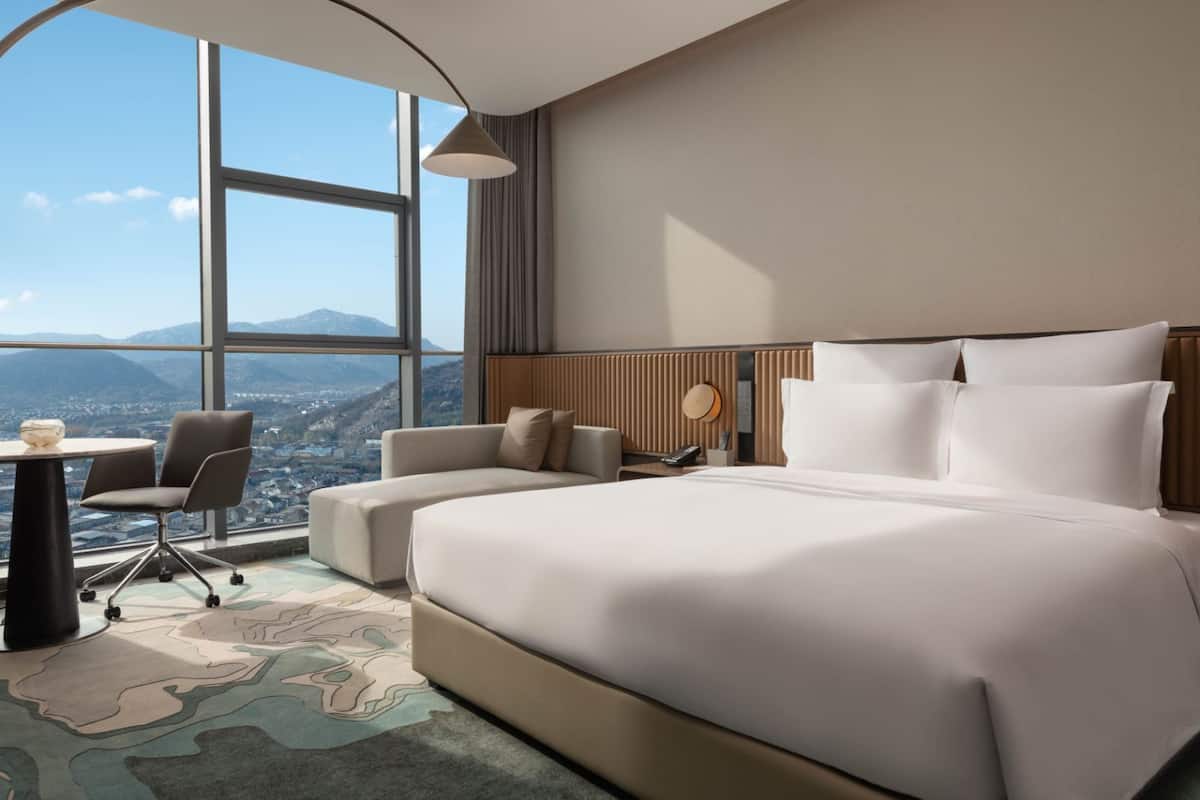 Ein modernes Hotelzimmer mit einem großen Bett, einer Couch, einem Schreibtisch und Blick auf eine Stadtlandschaft durch große Fenster.