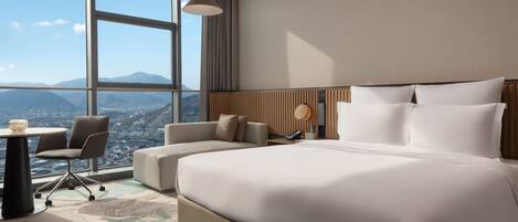 Ett modernt hotellrum med en stor säng, en soffa, ett skrivbord och utsikt över stadsbilden genom stora fönster.