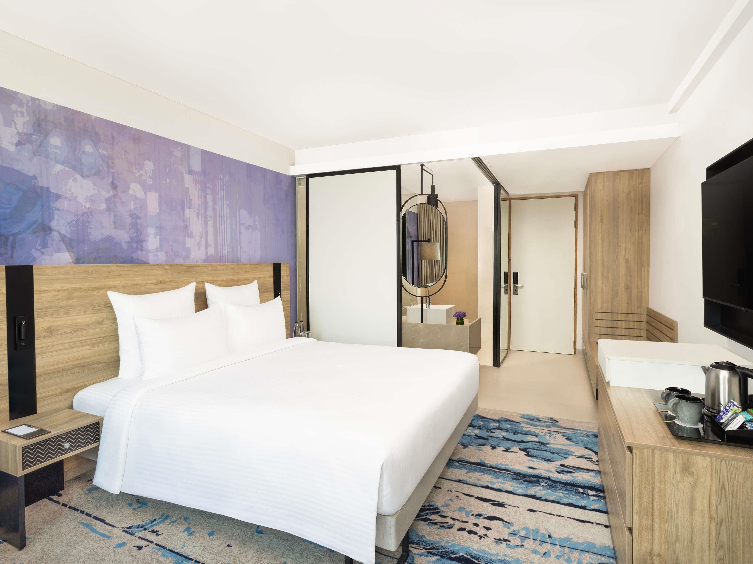 A modern hotel room with a large bed, a wooden headboard, a flat-screen TV, a desk with a coffee maker, and a mirror.