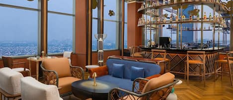 A high-rise bar with a panoramic city view, featuring modern furniture and a central bar area.