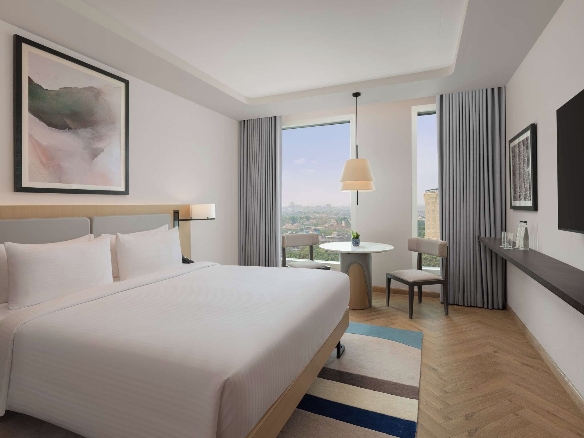 A hotel room with a large bed, a desk, a chair, a small table, and a view of the city.