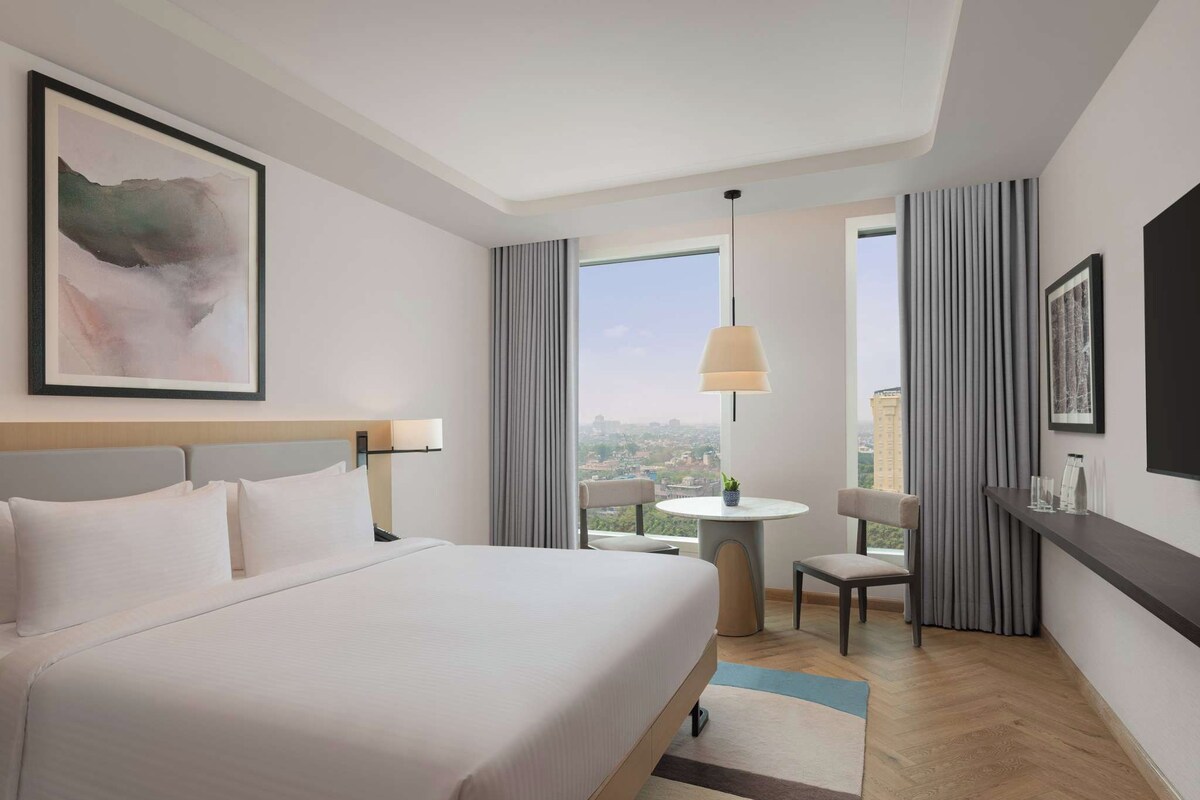 A hotel room with a large bed, a desk, a chair, a small table, and a view of the city.