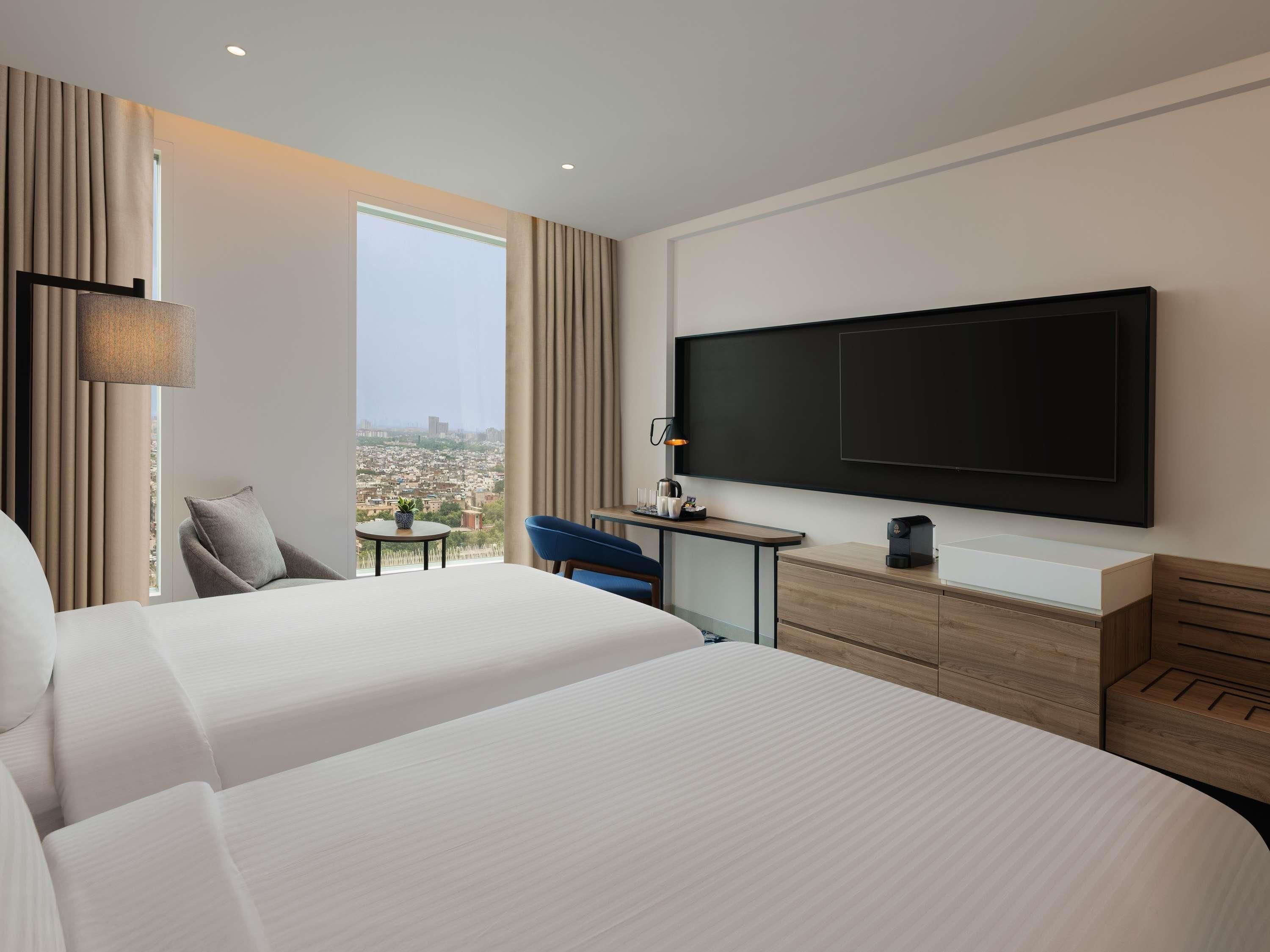 A hotel room with two beds, a TV, a desk with a lamp, and a view of the city.