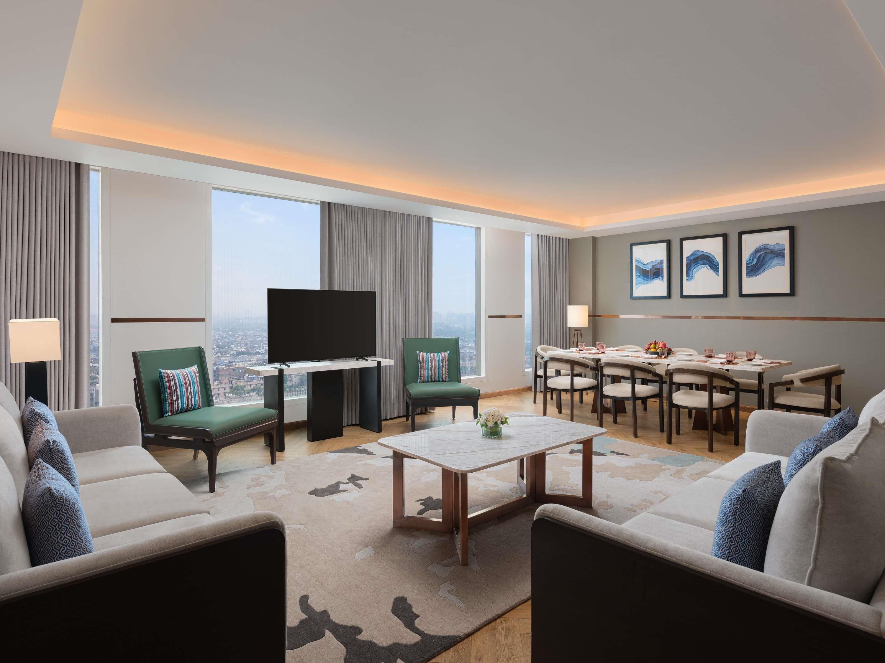 A modern hotel room with a flat-screen TV, green armchairs, a dining table, and a city view.