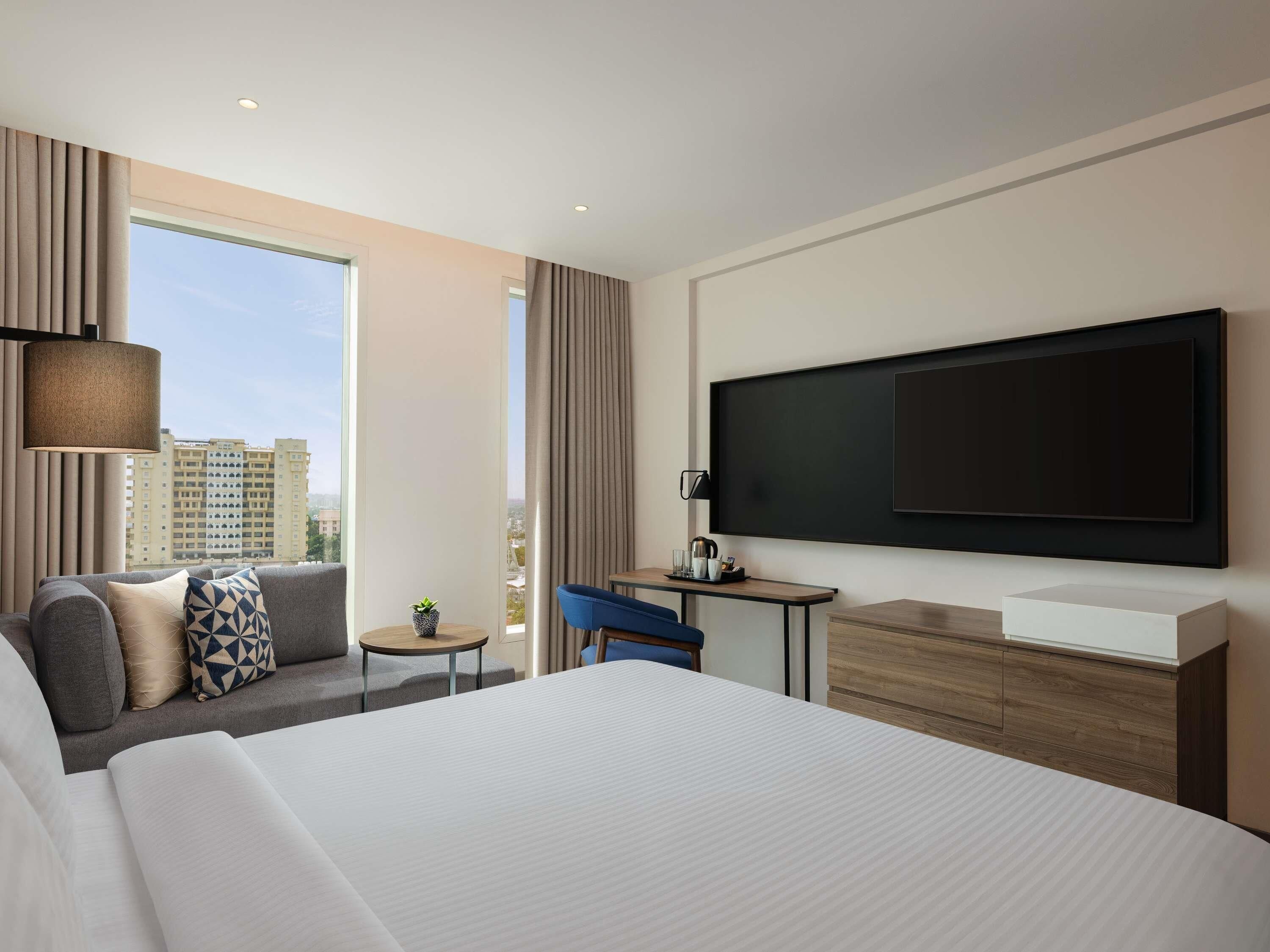 A modern hotel room with a large bed, a sofa, a small table, a TV, and a window with a city view.