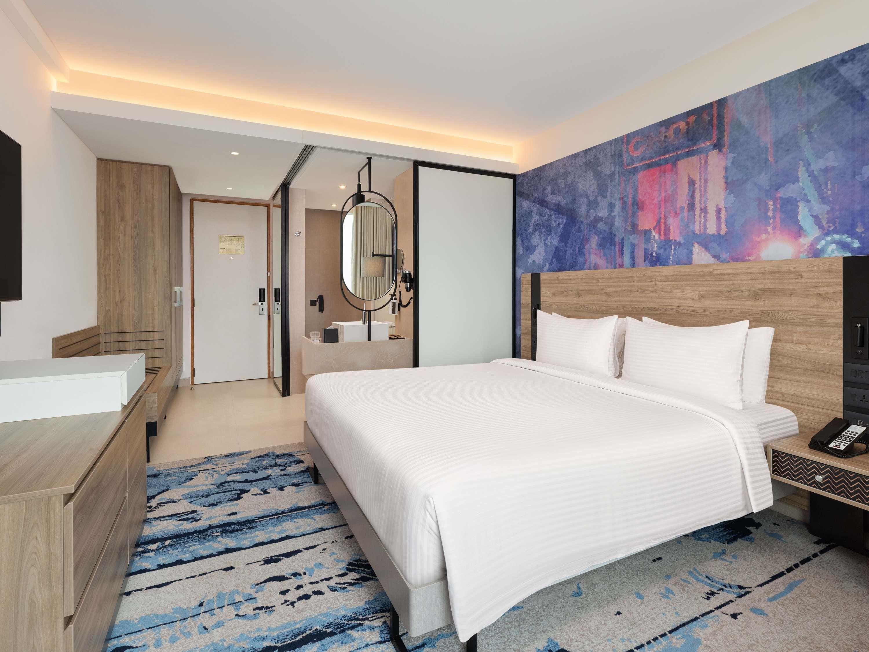 A modern hotel room with a large bed, a wooden headboard, a bedside table with a phone, a mirror, and a bathroom with a wooden vanity.