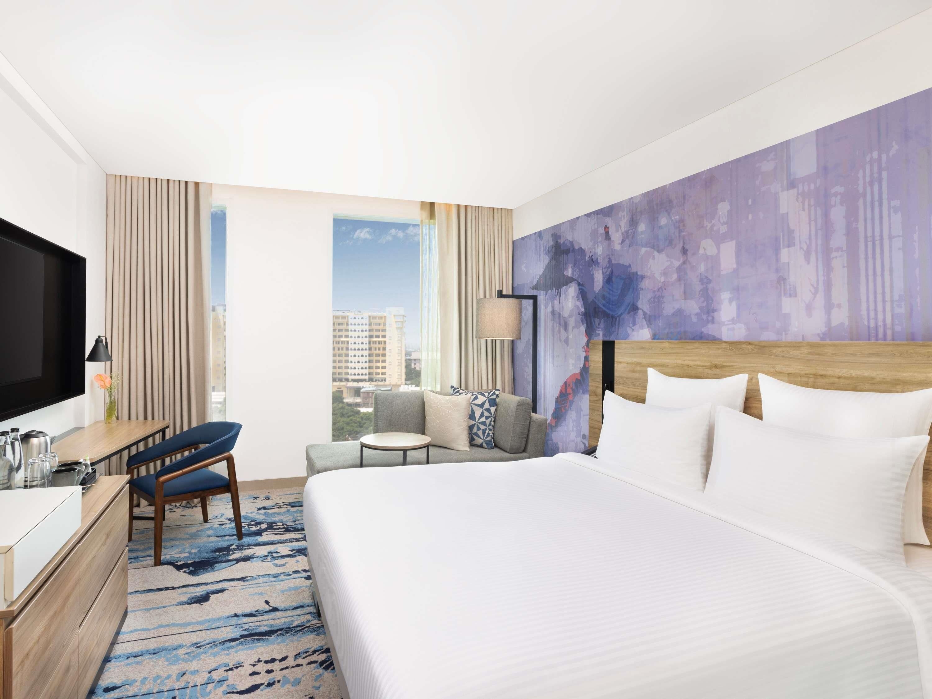 A hotel room with a bed, a desk with a chair, a sofa, a TV, and a window with a city view.