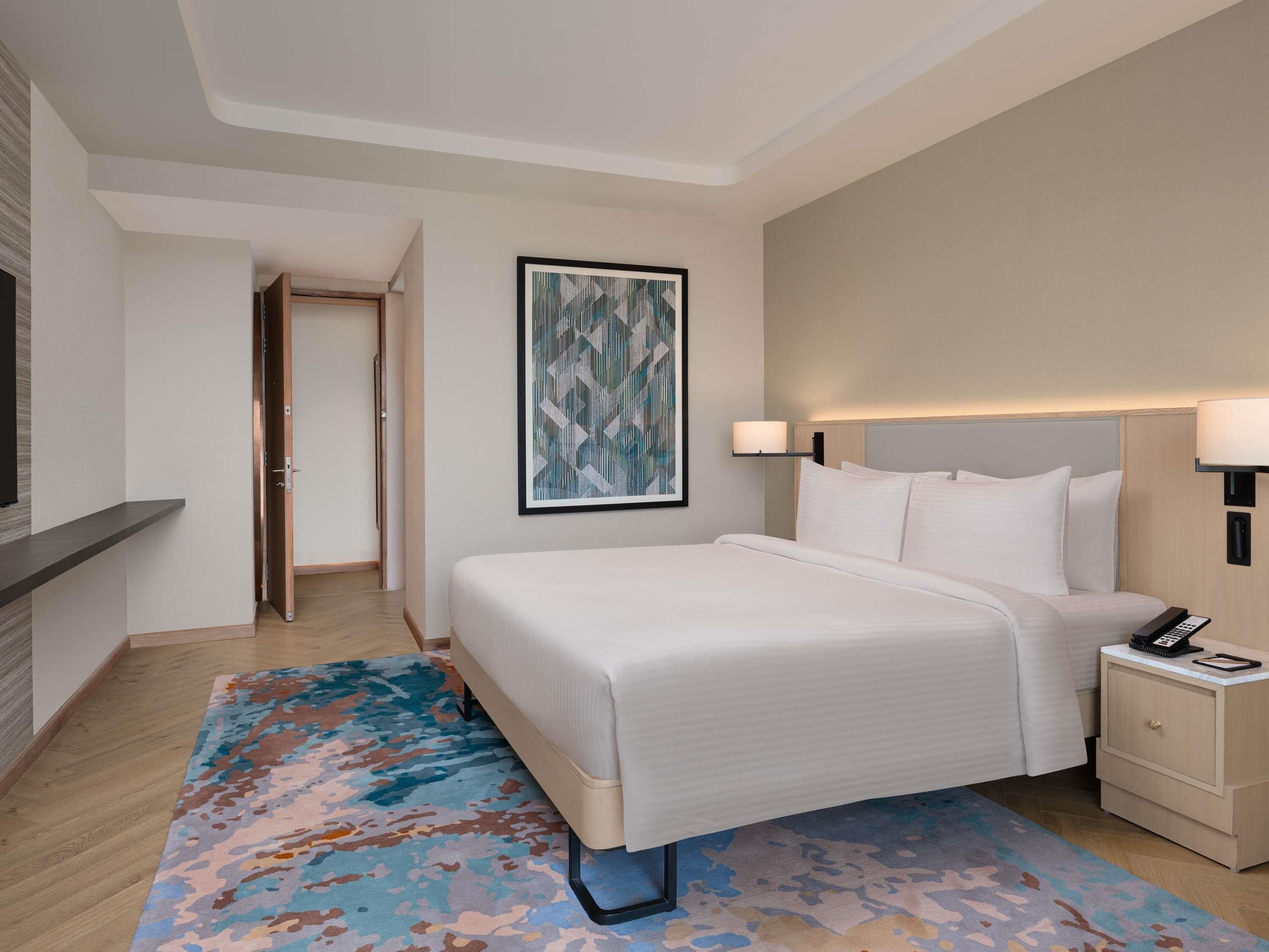 A hotel room with a bed, bedside table, lamp, and a painting on the wall.