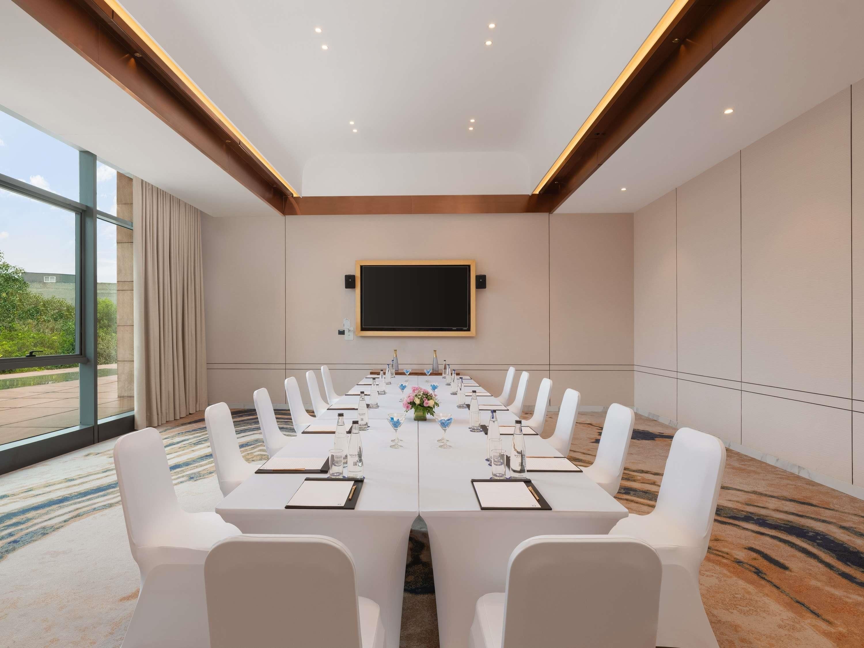A conference room with a long table, white chairs, a mounted screen, and a view through large windows.