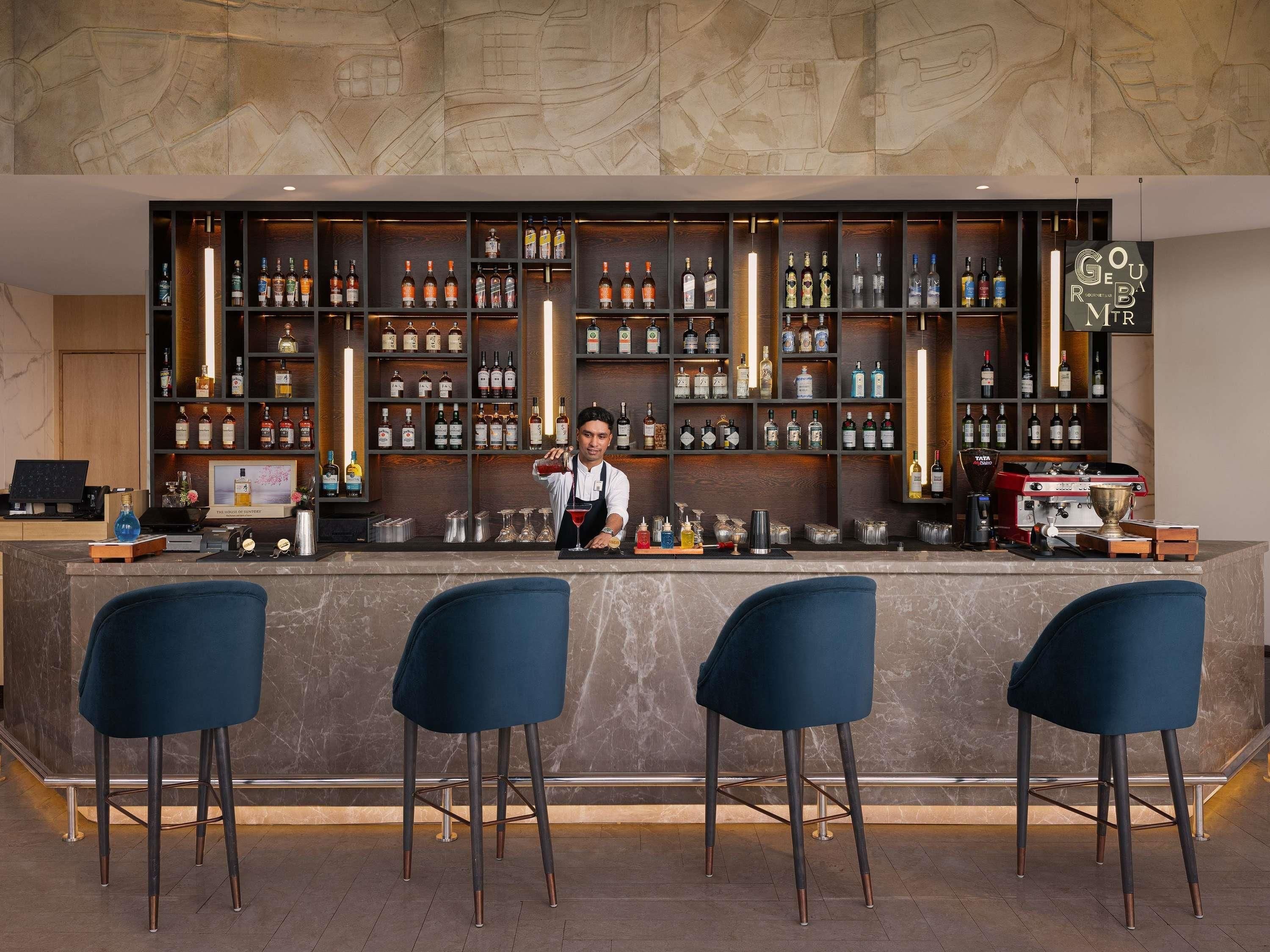 Een bar met een barman, meerdere planken vol drankflessen, een koffiemachine en drie blauwe barkrukken.