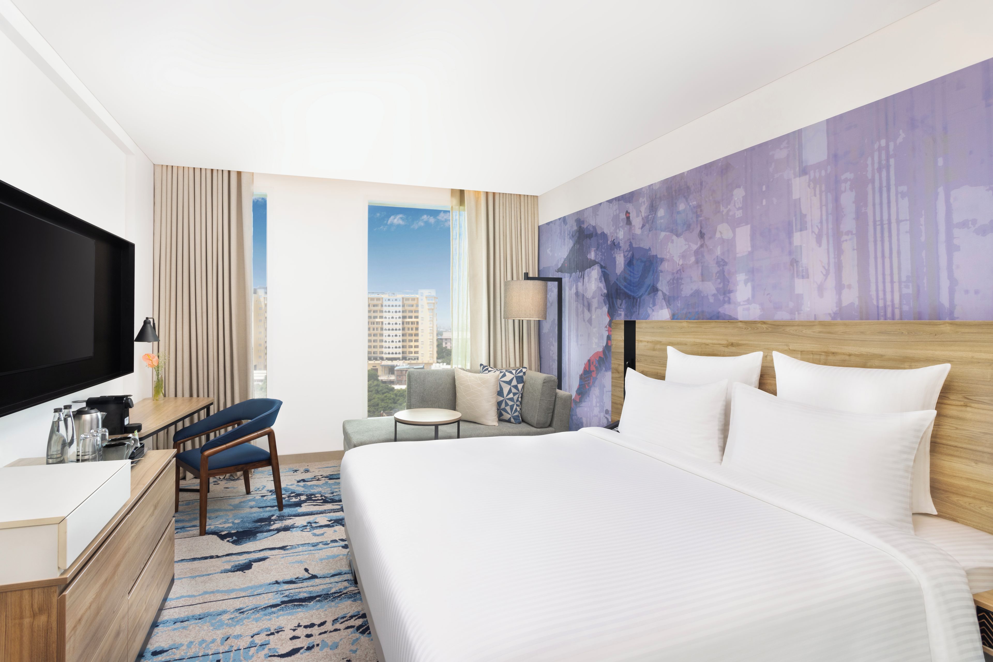 A hotel room with a large bed, a desk with a flat-screen TV, a chair, a small table, and a view of the city through the window.