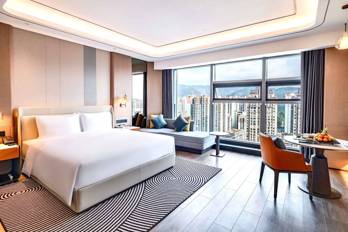 Habitació d'hotel moderna amb un llit gran, un sofà, una taula de dinar i vista a la ciutat.