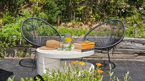 A round table with a white top, surrounded by two black wire chairs. On the table, there is a straw hat, a book titled 'Book of Colours', and a clear glass pitcher with a yellow flower.