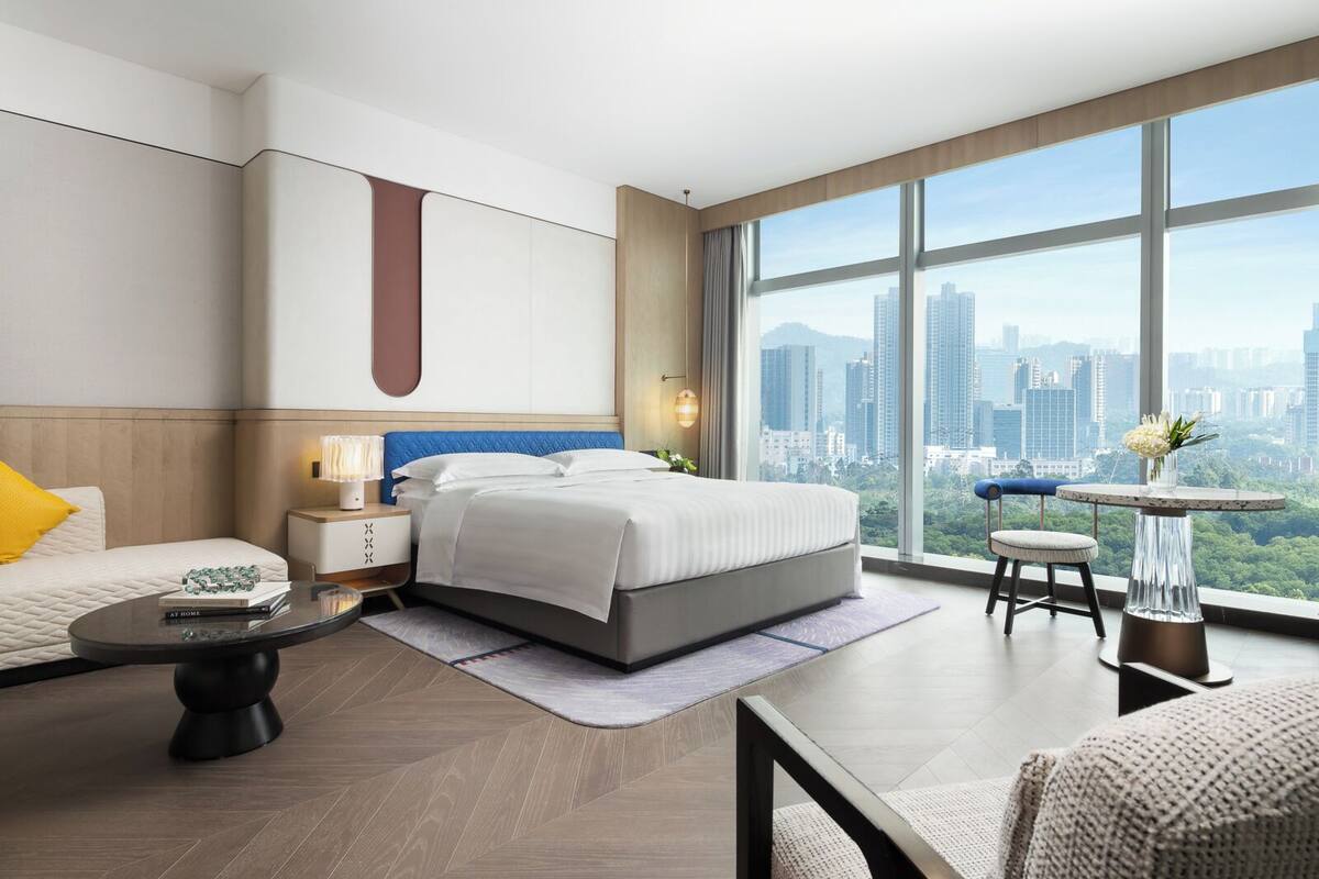 A modern hotel room with a large bed, a sitting area with a table, and a view of the cityscape.