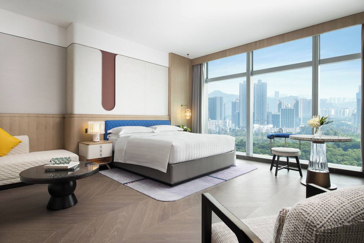 A modern hotel room with a large bed, a sitting area with a sofa and armchair, a small round table, and a view of the city through large windows.