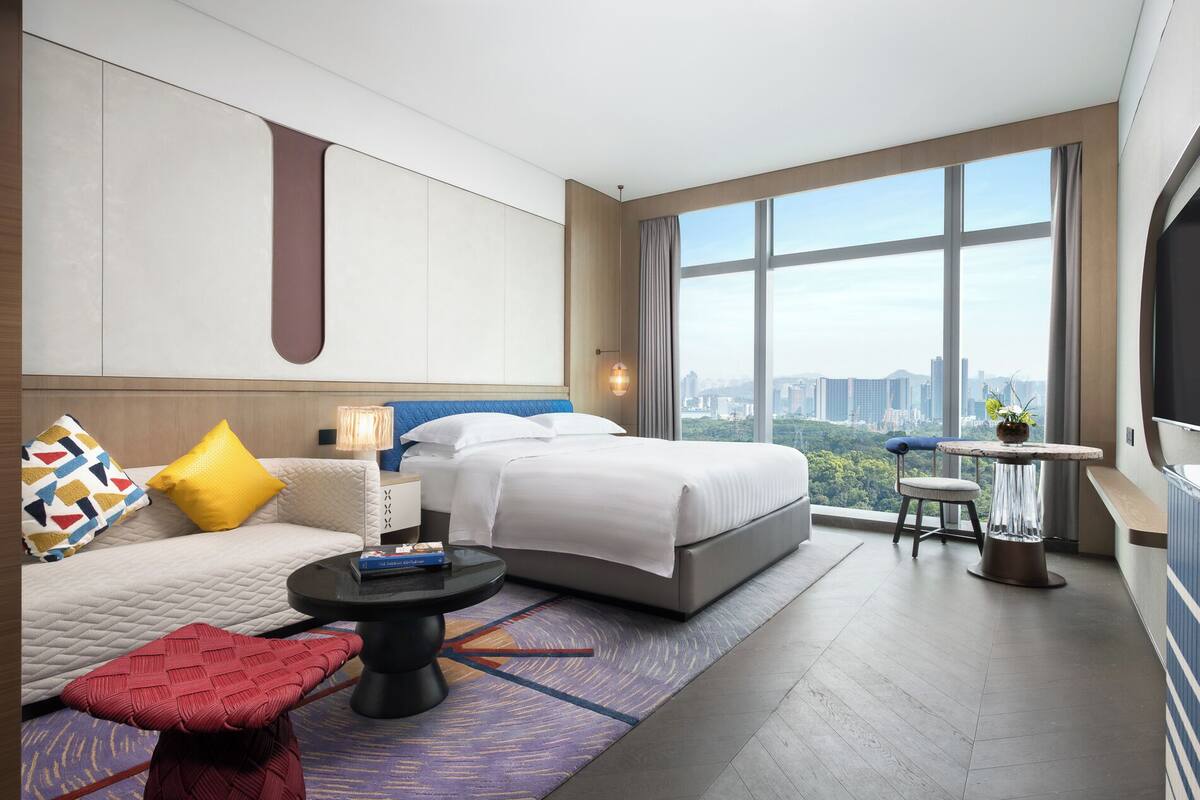 A modern hotel room with a large bed, a sitting area with a sofa and armchair, a small round table, and a large window offering a city view.