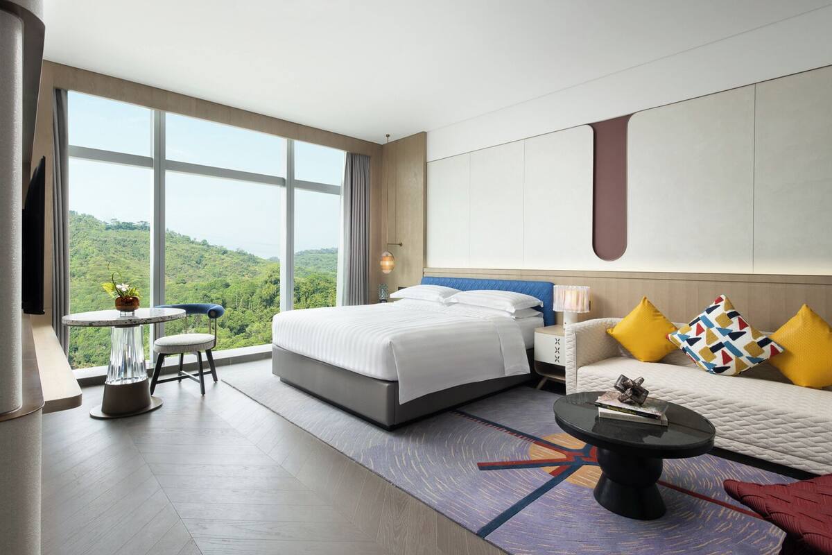 A modern hotel room with a large bed, a sofa, a small table, and a view of the outdoors.