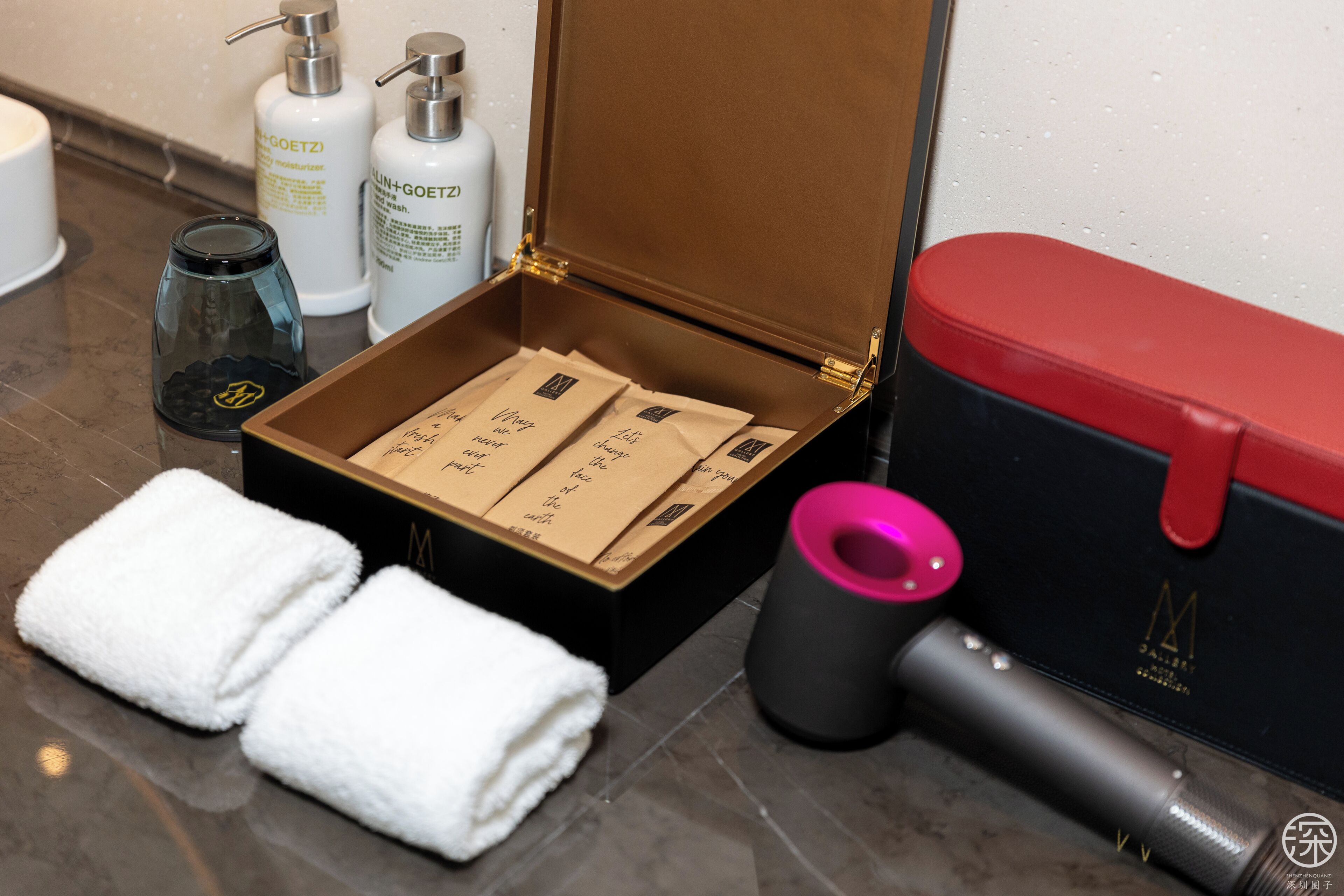 A bathroom with a wooden box containing toiletries, a black and red container, a pink and black cylindrical object, and two white towels.