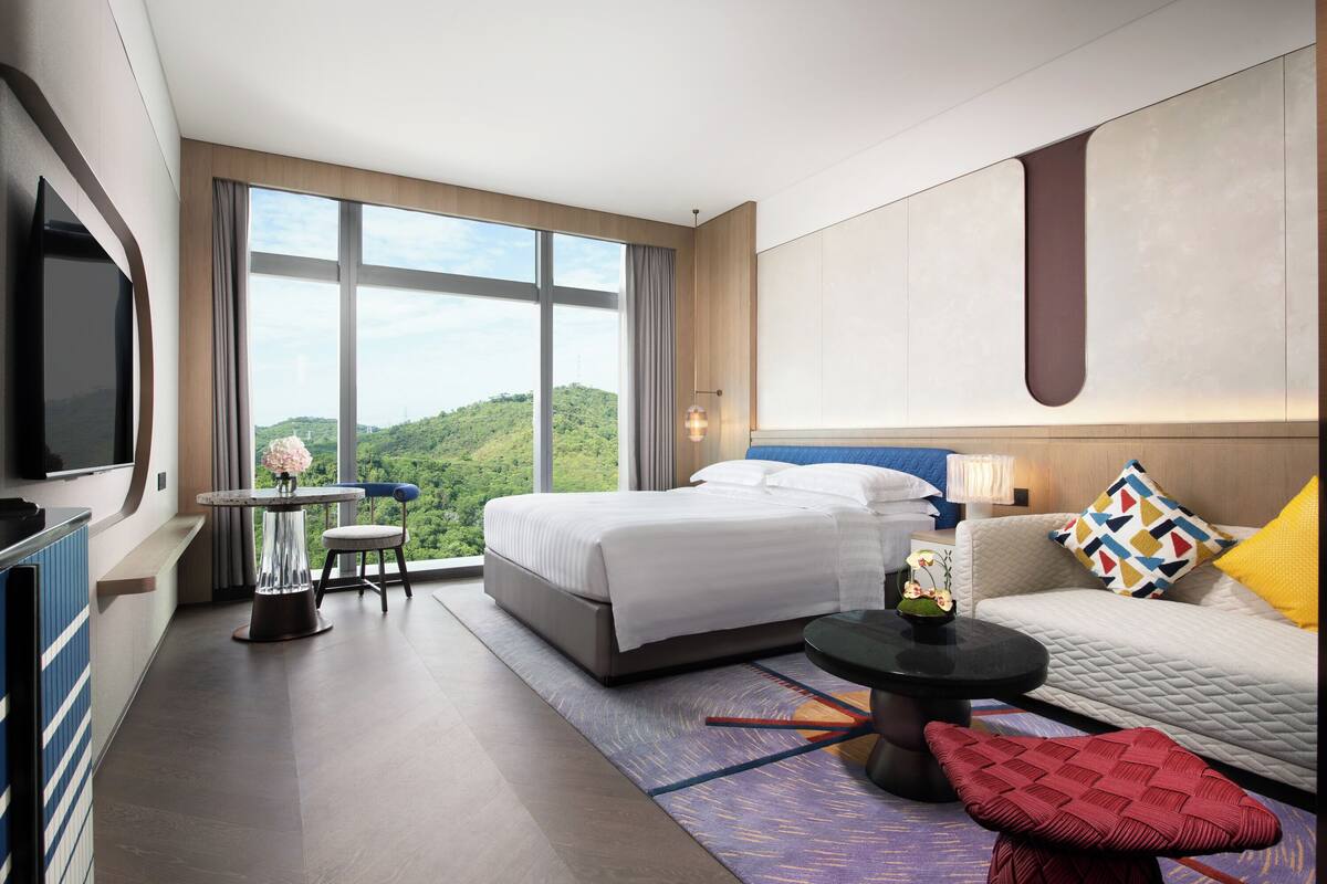 A modern hotel room with a large bed, a sofa, a TV, and a view of the outdoors.