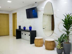 A modern lobby with a marble wall, a flat-screen TV, a large mirror, a console table with decorative vases, two wooden stools, and potted plants. - Ibis Styles Foz Do Iguaçu (Foz do Iguaçu)