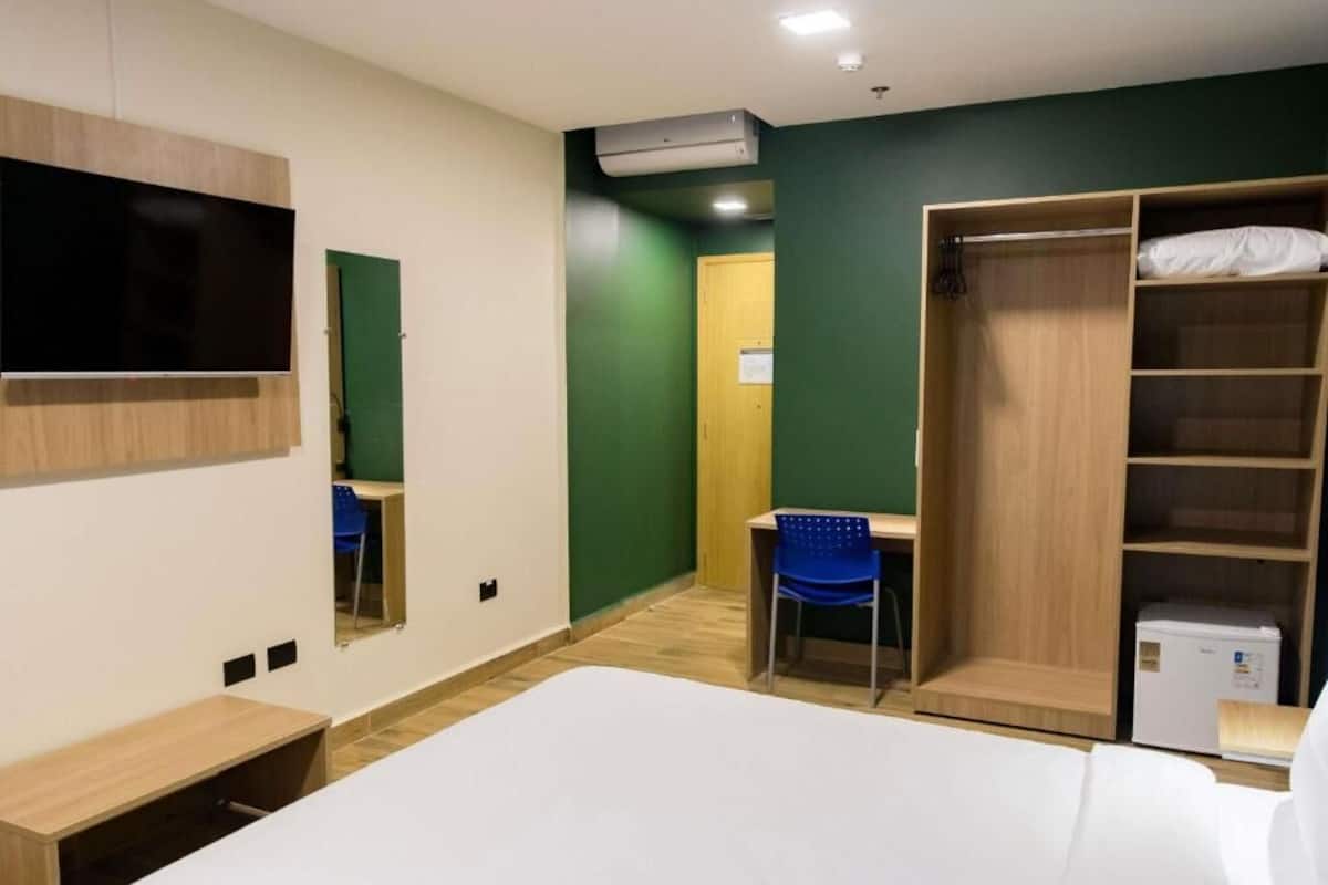 Ein Hotelzimmer mit einem Bett, einem Schreibtisch, einem Stuhl, einem Kleiderschrank, einem Fernseher und einer Klimaanlage.