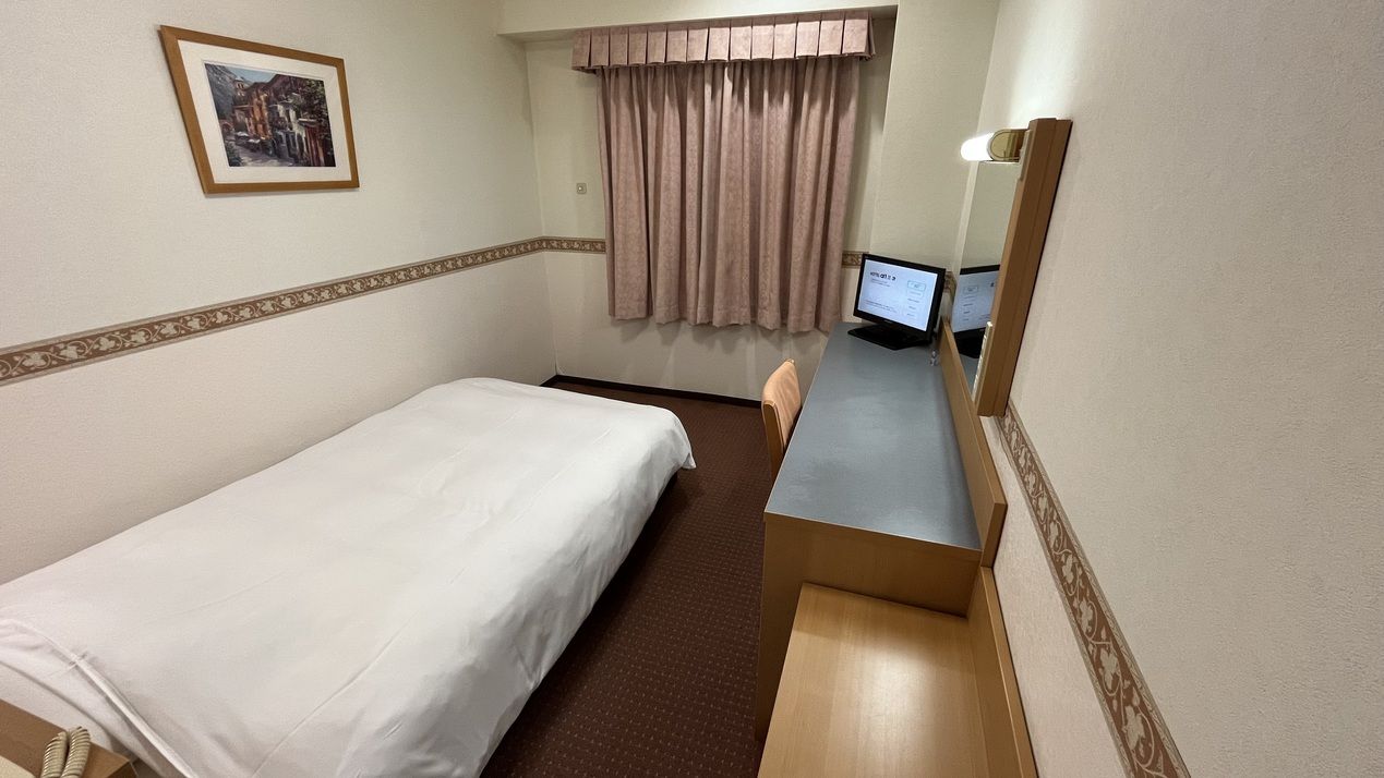 A hotel room with a single bed, a desk with a computer, a chair, and a window with curtains.