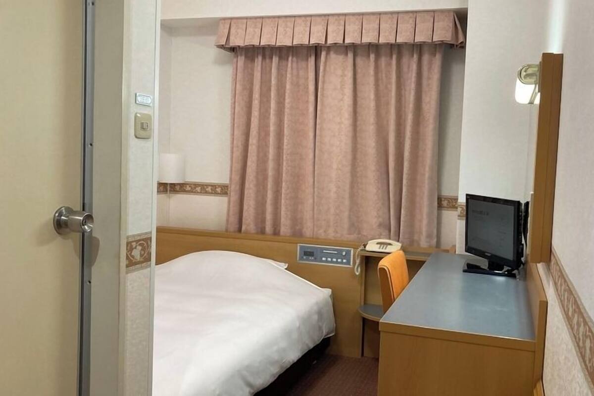A hotel room with a bed, a desk with a computer, a chair, a telephone, and a window with curtains.