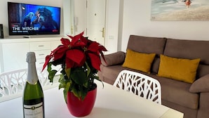 Un salón con un televisor que muestra 'The Witcher', una planta en maceta roja, una botella de Cava Jaume Serra y un sofá marrón con almohadones mostaza.