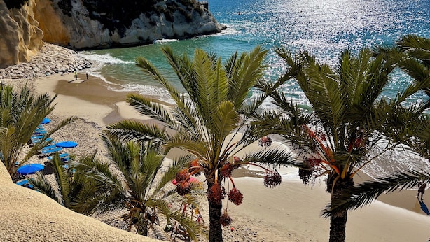 Una playa con aguas azules cristalinas, un acantilado rocoso y palmeras.