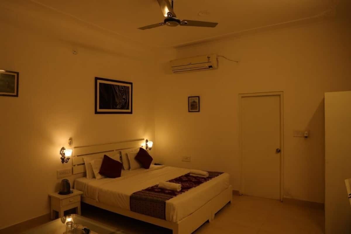 A hotel room with a bed, bedside table, nightstand, wall-mounted air conditioning unit, ceiling fan, and a door.