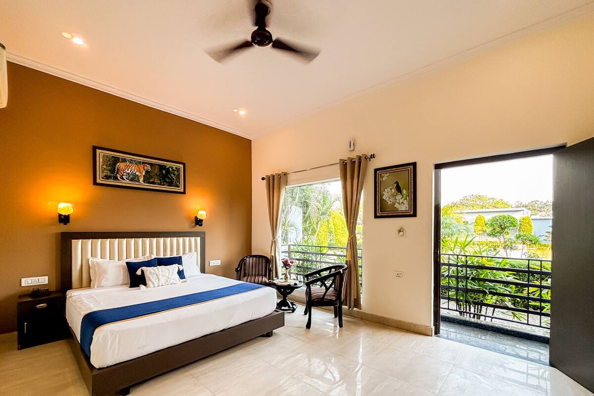 A hotel room with a large bed, a ceiling fan, a nightstand, a table with a chair, and a balcony with a view of greenery.