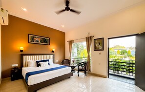 Habitación de hotel con una cama grande, un ventilador de techo, una mesita de noche, una mesa con una silla y un balcón con vistas a zonas verdes.