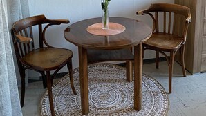 Un comedor con una mesa redonda de madera, dos sillas de madera, un jarrón con flores, un tapete estampado y un cuadro en la pared.