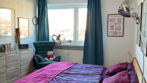 Un dormitorio con cama, cómoda, silla, ventana con cortinas, reloj y un cuadro en la pared.