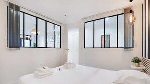A modern hotel room with a large bed, two windows with black frames, a bedside table with a lamp, and a small potted plant. - Super appartment-2BR/6p-Bastille (Paris)