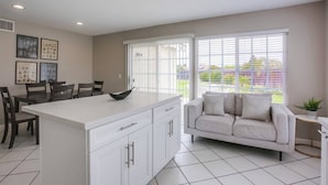 A modern kitchen with a central island, dining area with chairs, a sofa, and large windows with blinds.