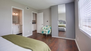 A bedroom with a bed, a vanity with a mirror, a chair, and a large mirror on the wall.