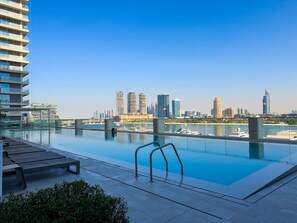 A rooftop infinity pool with a city skyline view, lounge chairs, and a clear sky.
