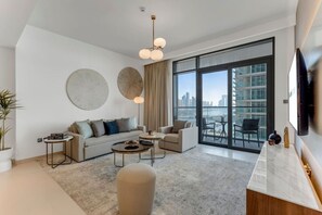 A modern living room with a sofa, two armchairs, a coffee table, and a TV. There is a large window with a city view. - Coastal Chic 1 Bedroom Apartment w private beach (Dubai)