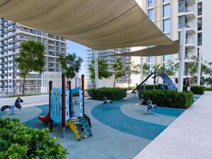 Ein Spielplatz mit verschiedenen Spielgeräten, umgeben von einem überdachten Bereich und einem Gebäude im Hintergrund.