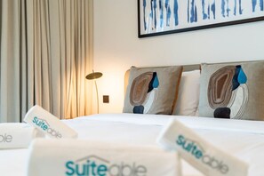 Ein Hotelzimmer mit Bett, Kissen, Vorhängen, einer Wandleuchte und einem gerahmten Kunstwerk.