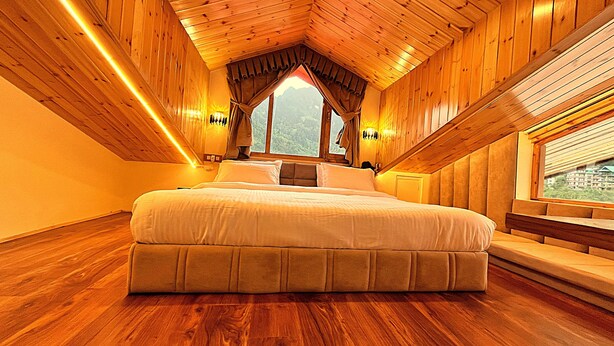 A bedroom with a large bed, wooden walls, a sloped ceiling, and a window with a view of mountains. - Wander Thrives Inn By Ads (Manali)