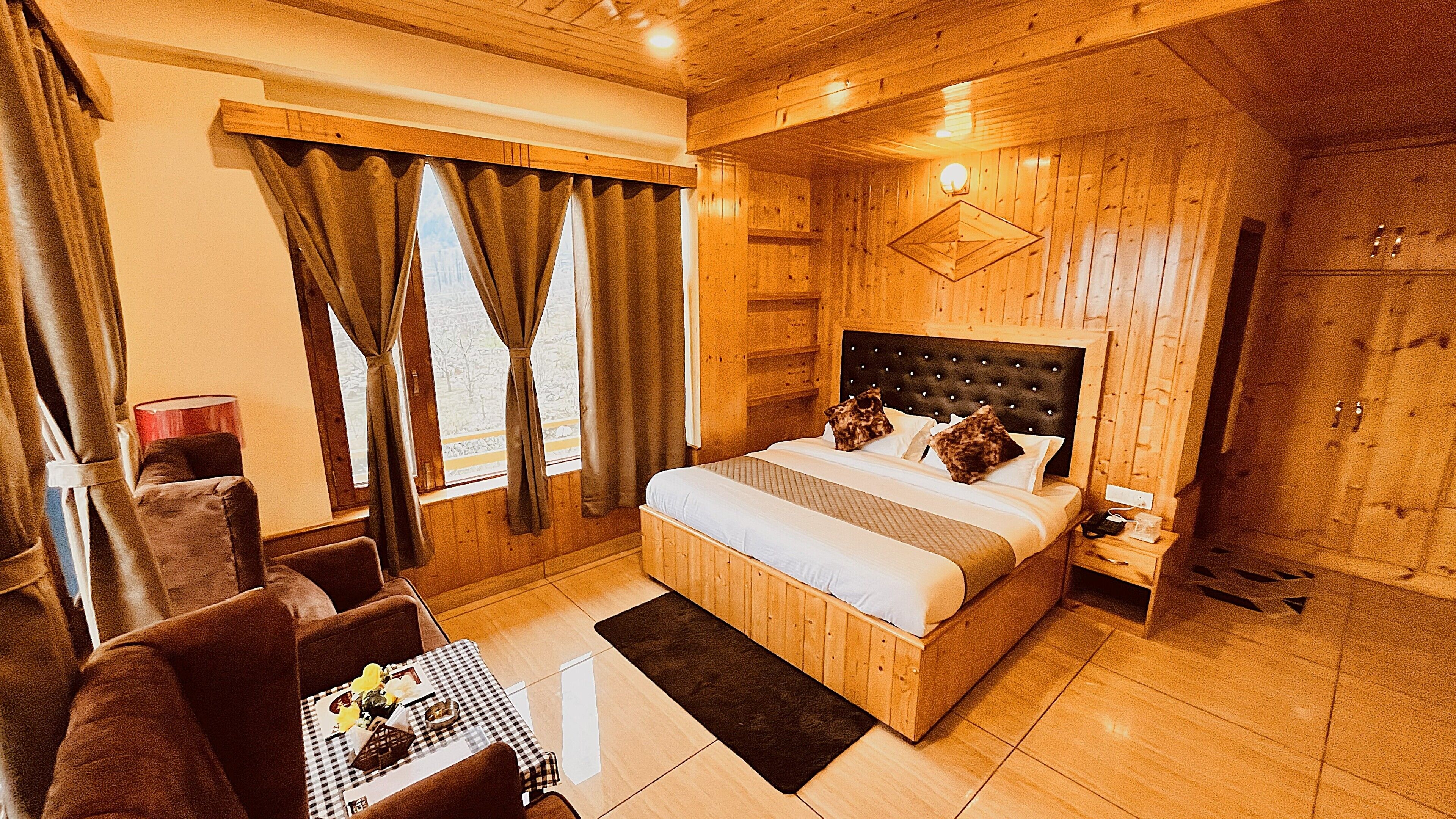A wooden room with a bed, a chair, a table with a checkered tablecloth, and curtains on the windows.
