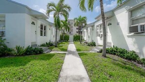 A pathway leading to a row of white buildings with palm trees and a clear sky.